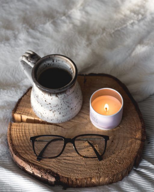 A mug of coffee, a lit candle, and a pair of eyeglasses sit on a round piece of wood on a bed with blankets in the background.