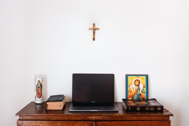 an open laptop sitting on a desk surrounded by a bible, prayer books, and religious icons.