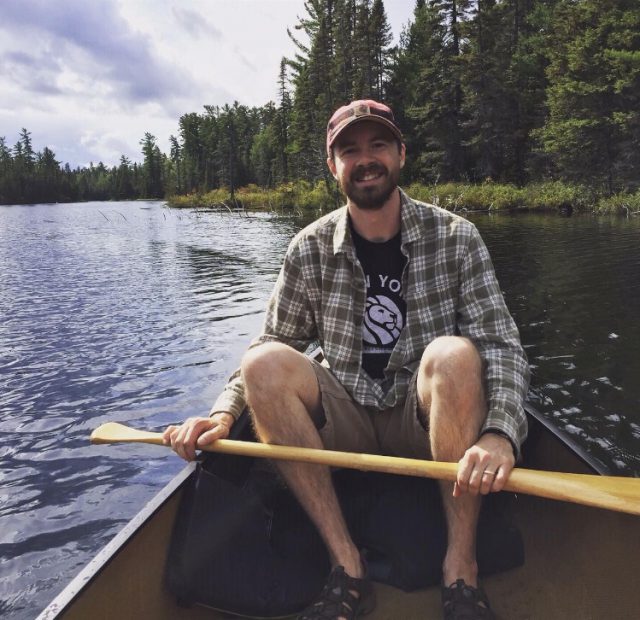 Mike, the author, sits in a canoe on a lake. He holds a paddle, Is wearing shorts and has a beard. He smiles at the camera