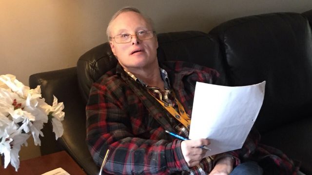 A man with Down syndrome and grey hair is sitting on a couch and looks up at the camera