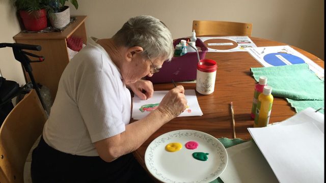 An older man sits at a kitchen table with art supplies spread around him as he works on a painting.