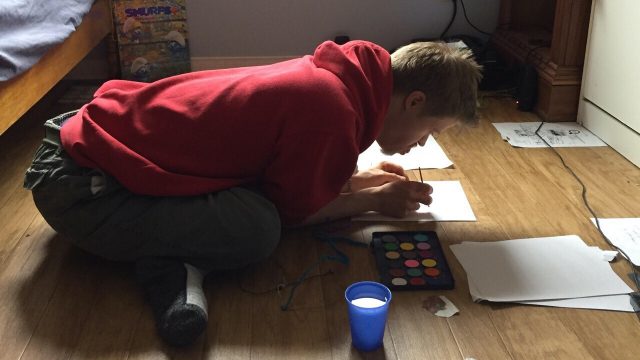 A younger man with Down syndrome leans over on the floor of his bedroom as he paints a picture on a sheet of paper
