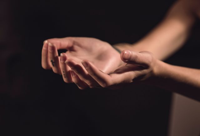 two hands, held palm up as if in prayer. A small tattoo of a cross is visible on one finger.