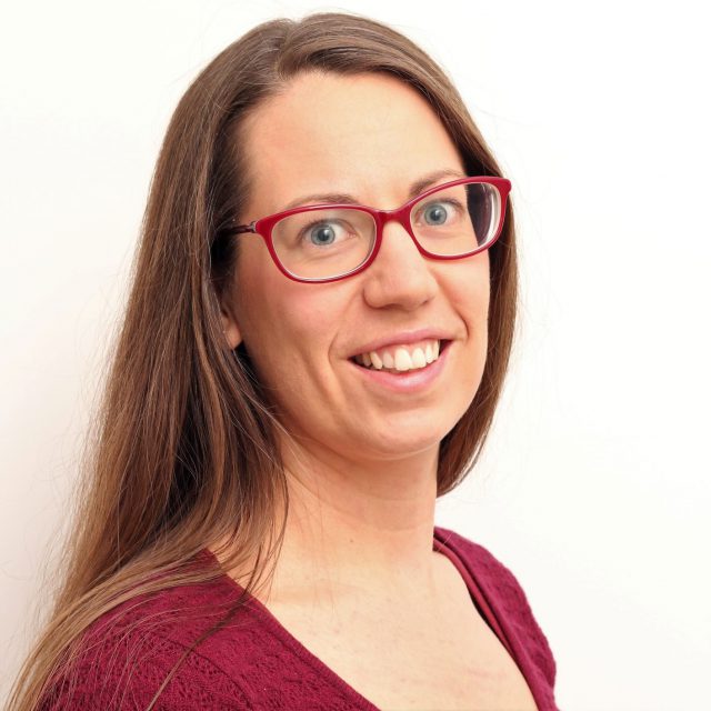 Emma smiles at the camera, wearing red glasses and a dark red tshirt.