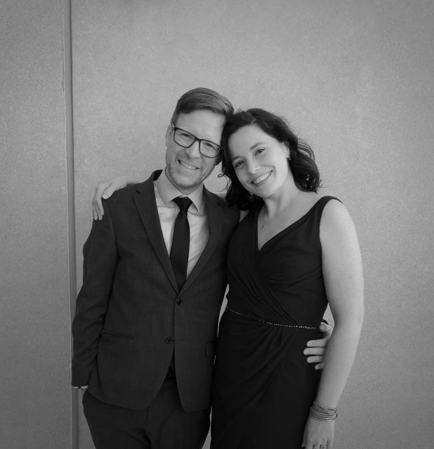 Carlo is wearing a suit, dark-framed glasses and is smiling while he has his arm around his wife Allyson who stands next to him. She is wearing a black dress and is also smiling.