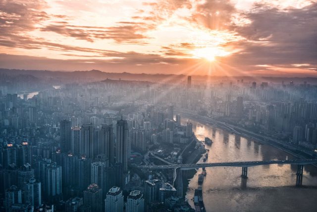 The sun rises over a city filled with skyscrapers, shining long beams of light onto various buildings.