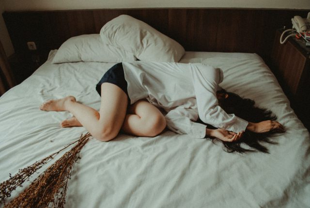 a woman lies on a bed, curled up in a fetal position, with her arms covering her face.