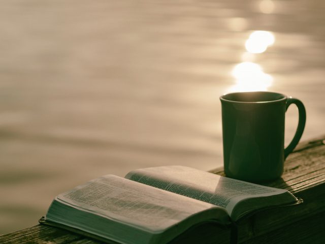 A bible and mig sit on a wooden railing beside a lake at sunset.