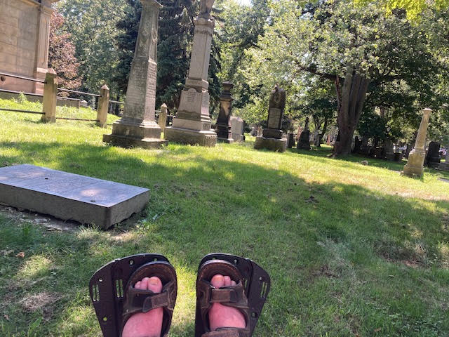 Dion's feet, on his wheelchair foot rests, are visible in the foreground of the photo. In the backgroung are tombstones and trees.