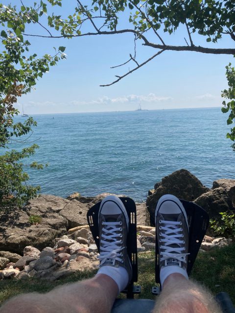 Dion's feet, wearing converse sneakers, are visible in the foreground, resting on his wheelchair foot rests. in the background is a rocky shore and then a wide expanse of lake.