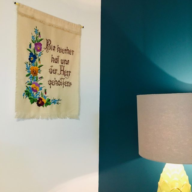 An embroidered wall hanging that says "Bis hierher hat uns der Herr geholfen!" is hanging on a white wall, beside a teal wall. A lamp stands nearby.