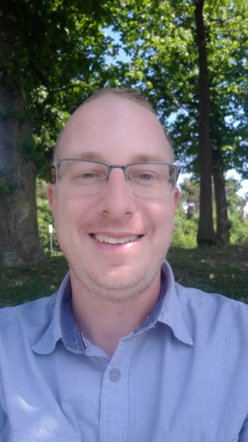 Steve has light skin, glasses, and is smiling directly into the camera. He stand outside and there are trees behind him.