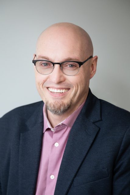 Dwayne is a white man who is bald and has a goatee. He wears a dark suit, and glasses and he is smiling at the camera.