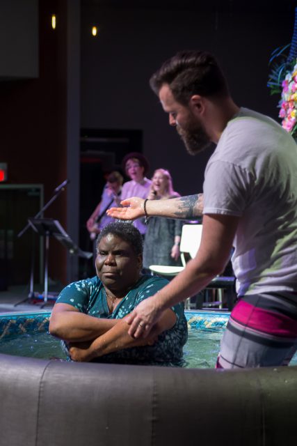 A Black woman kneels in a baptismal tank. A white man stands beside her preparing to baptize her.