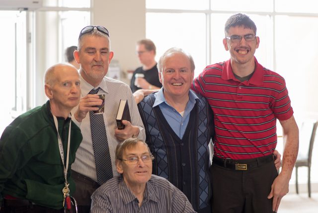 A group of white men, of varying ages, with and without visible disabilities stand together in a church lobby. Most are smiling at the camera and some have their arms around each other.