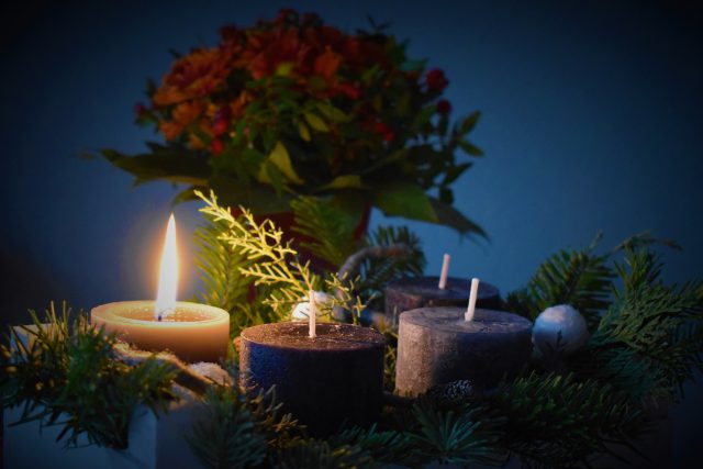 One candle is list beside three unlit candles. There is greenery around the candles and a bouquet of flowers in the background.