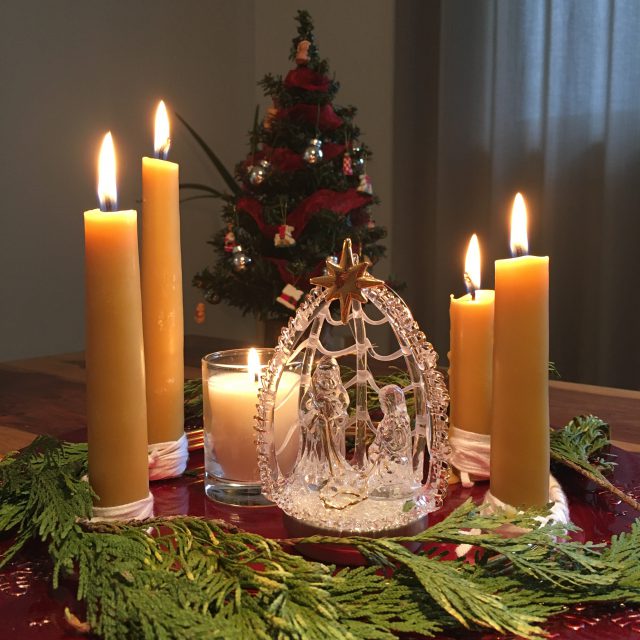 An advent wreath sits on a table with a small glass nativity set in the centre all five candles are lit . There is a small Christmas tree in the background