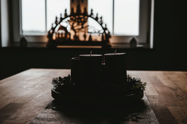 An advent wreath sits on a table. The candles are unlit and it is in shadow. In the background is a nativity scene.