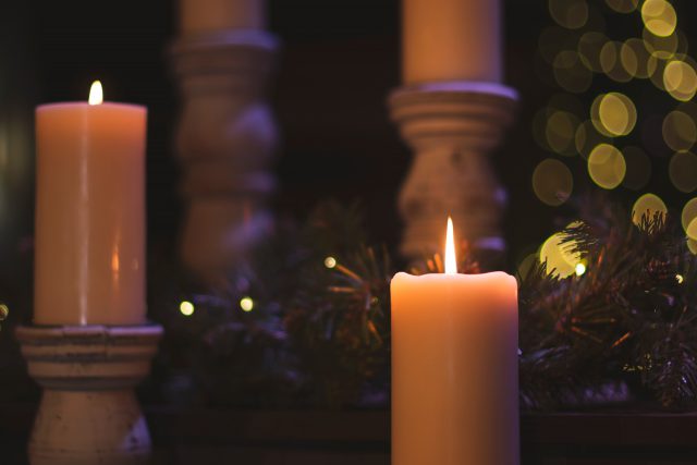 Two lit candles can be seen in front of greenery, and two more candles in the background.