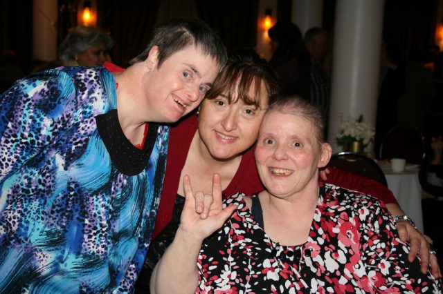Three white women lean close together and smile at the camera. The woman on the left has down syndrome, and is standingthe woman on the right is making the "I Love You" sign to the camera and is seated. The woman in the middle has her arms around both other women and is drawing all three together.