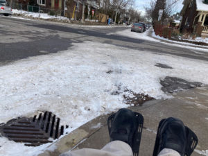 Dion's outstretched legs are visible as he takes a photo from his position sitting in his wheelchair. The sidewalk in front of him is fairly clear, but beyond the curb there is lots of snow and a drain in the street visible.There is a curb cut in front of him, leading down to the street.
