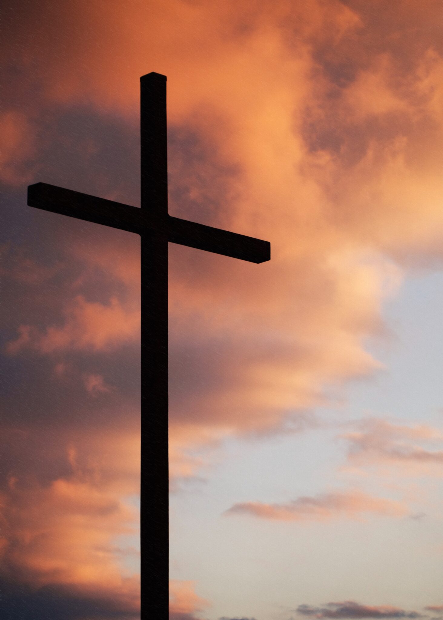 A cross stands in silhouette in front of a sky at sunset. There are orange and pink clouds in front of a blue sky.